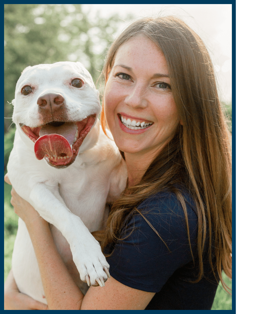 Photo of a brown haired woman smiling and holding a happy white bully-breed dog taken outdoors on a sunny day